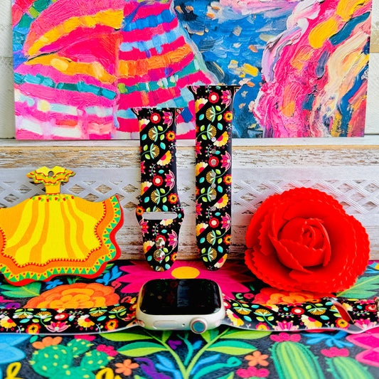 Bloom Bonita silicone Apple Watch band with Mexican folk art florals in coral, red, yellow, teal, and pink on a black base, shown displayed against a vibrant Mexican folk art backdrop with rose and dress accents.