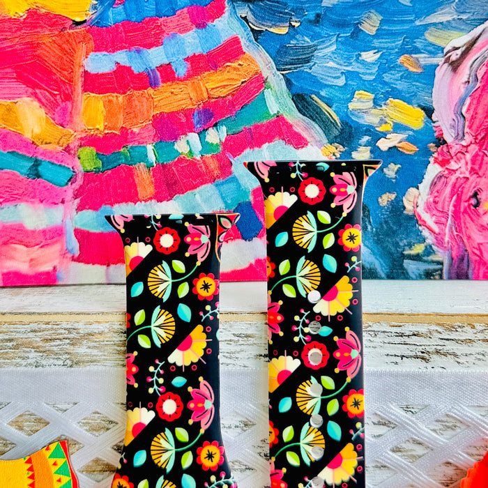 Bloom Bonita silicone Apple Watch band with Mexican folk art florals in coral, red, yellow, teal, and pink on a black base, shown displayed against a vibrant Mexican folk art backdrop with rose and dress accents.