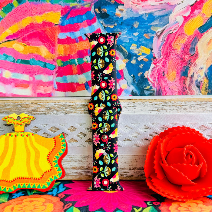 Bloom Bonita silicone Apple Watch band with Mexican folk art florals in coral, red, yellow, teal, and pink on a black base, shown displayed against a vibrant Mexican folk art backdrop with rose and dress accents.