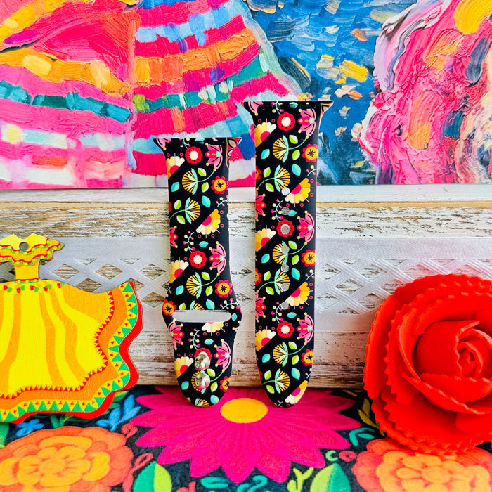 Bloom Bonita silicone Apple Watch band with Mexican folk art florals in coral, red, yellow, teal, and pink on a black base, shown displayed against a vibrant Mexican folk art backdrop with rose and dress accents.