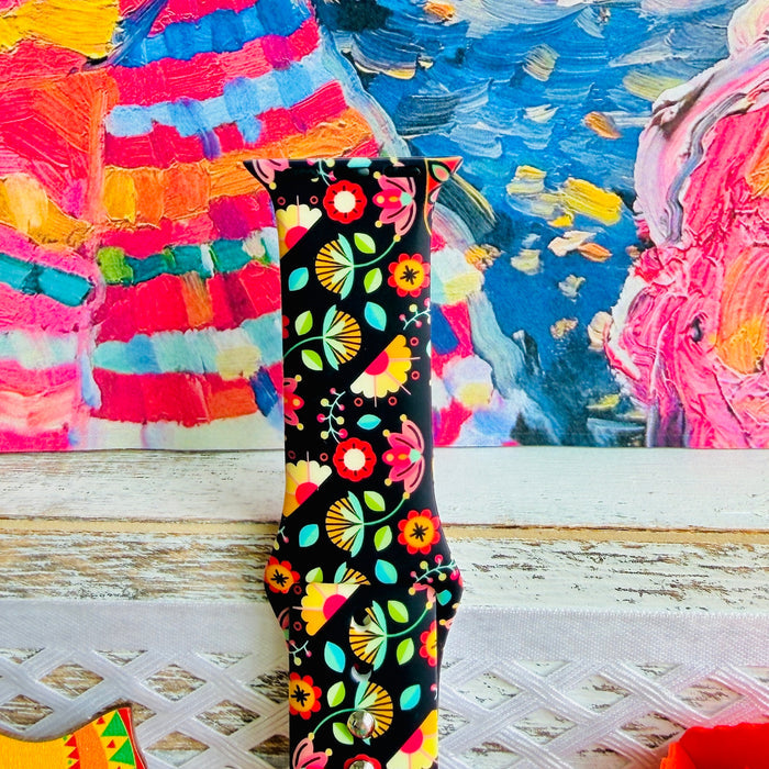Bloom Bonita silicone Apple Watch band with Mexican folk art florals in coral, red, yellow, teal, and pink on a black base, shown displayed against a vibrant Mexican folk art backdrop with rose and dress accents.
