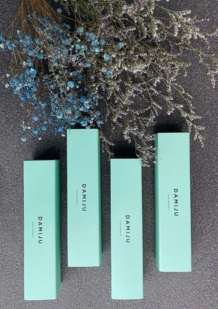 Four turquoise cosmetic boxes with 'DAMIU' branding on a dark surface with decorative plants.