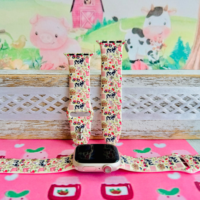 Farm Girl Cutie silicone Apple Watch band with dairy cows, red tractors, chickens, ducks, pink watering cans, strawberries, baskets, and roses on a cream base with pink gingham squares, shown displayed and worn on Apple Watch against a farmhouse country backdrop.