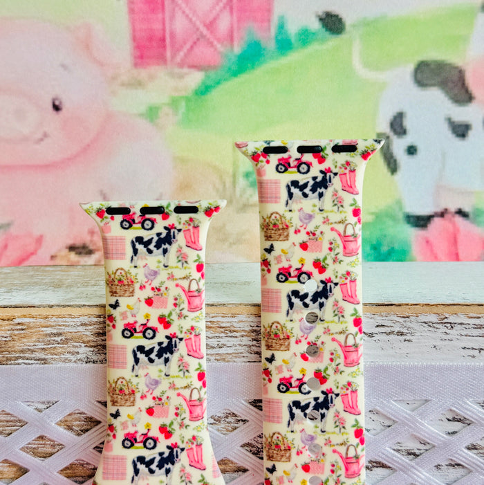 Farm Girl Cutie silicone Apple Watch band with dairy cows, red tractors, chickens, ducks, pink watering cans, strawberries, baskets, and roses on a cream base with pink gingham squares, shown displayed and worn on Apple Watch against a farmhouse country backdrop.