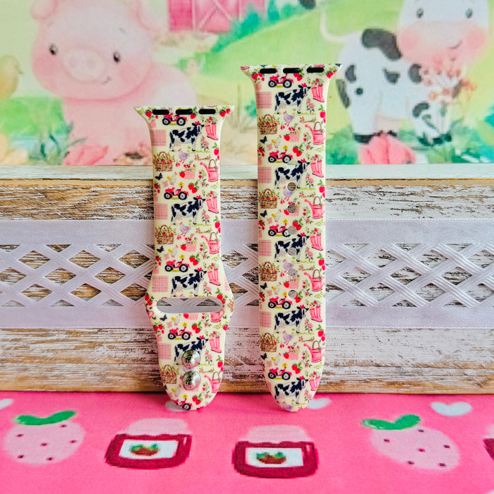 Farm Girl Cutie silicone Apple Watch band with dairy cows, red tractors, chickens, ducks, pink watering cans, strawberries, baskets, and roses on a cream base with pink gingham squares, shown displayed and worn on Apple Watch against a farmhouse country backdrop.