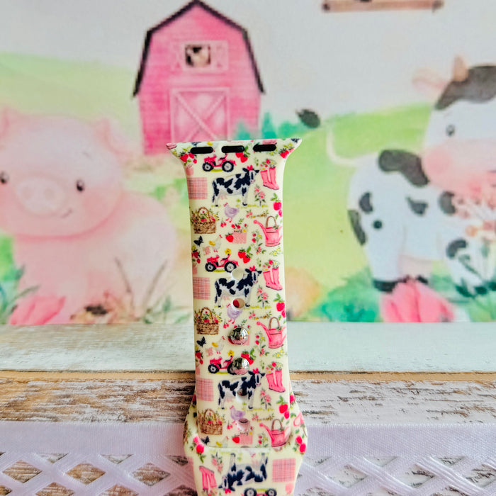 Farm Girl Cutie silicone Apple Watch band with dairy cows, red tractors, chickens, ducks, pink watering cans, strawberries, baskets, and roses on a cream base with pink gingham squares, shown displayed and worn on Apple Watch against a farmhouse country backdrop.
