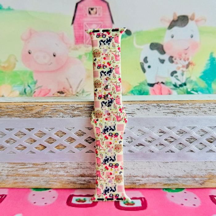 Farm Girl Cutie silicone Apple Watch band with dairy cows, red tractors, chickens, ducks, pink watering cans, strawberries, baskets, and roses on a cream base with pink gingham squares, shown displayed and worn on Apple Watch against a farmhouse country backdrop.