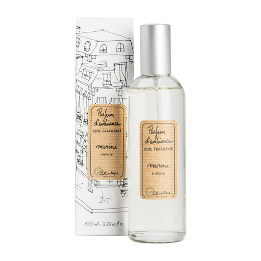 A clear glass room spray bottle with a label next to its packaging featuring a drawing of a house.