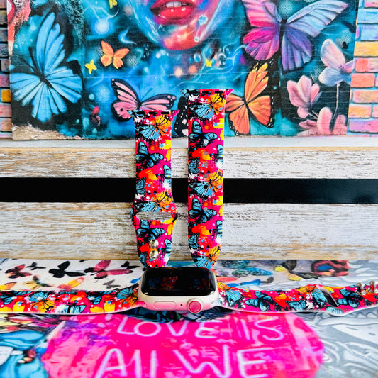 Rebel Wings silicone Apple Watch band with graffiti-style butterflies in blue, orange, yellow, and white over a hot pink and magenta paint-splatter background, shown displayed and worn on Apple Watch against a colorful street art butterfly backdrop.