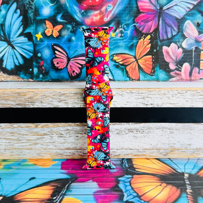 Rebel Wings silicone Apple Watch band with graffiti-style butterflies in blue, orange, yellow, and white over a hot pink and magenta paint-splatter background, shown displayed and worn on Apple Watch against a colorful street art butterfly backdrop.