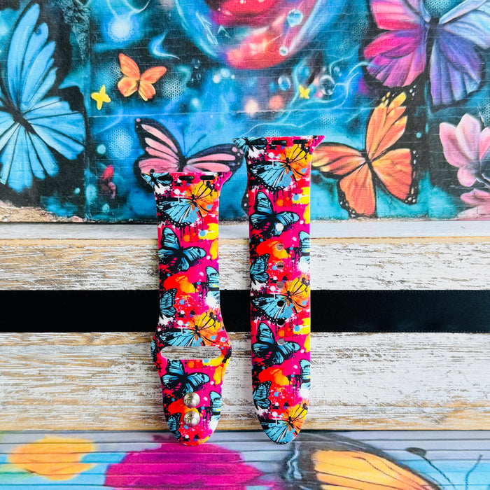 Rebel Wings silicone Apple Watch band with graffiti-style butterflies in blue, orange, yellow, and white over a hot pink and magenta paint-splatter background, shown displayed and worn on Apple Watch against a colorful street art butterfly backdrop.
