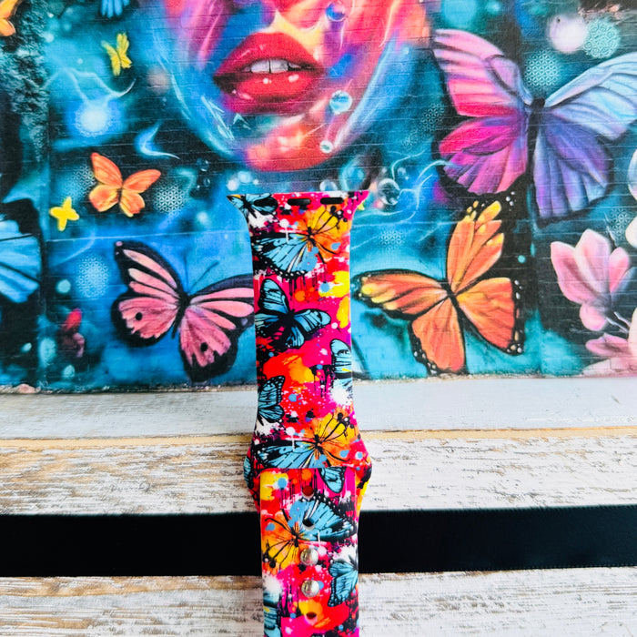 Rebel Wings silicone Apple Watch band with graffiti-style butterflies in blue, orange, yellow, and white over a hot pink and magenta paint-splatter background, shown displayed and worn on Apple Watch against a colorful street art butterfly backdrop.